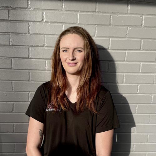 Person with long hair wearing a black T-shirt stands against a gray brick wall in sunlight.