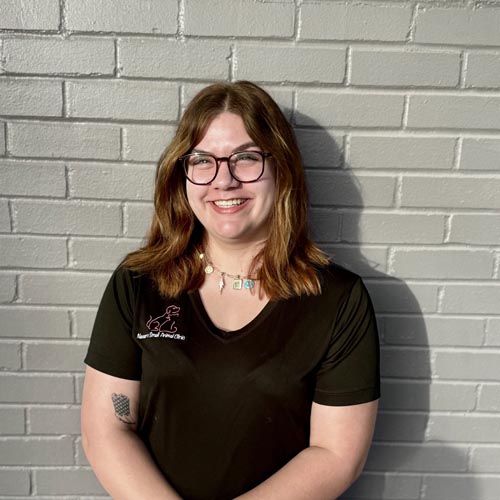 Person with brown hair and glasses smiling against a gray brick wall, wearing a black t-shirt and necklace.