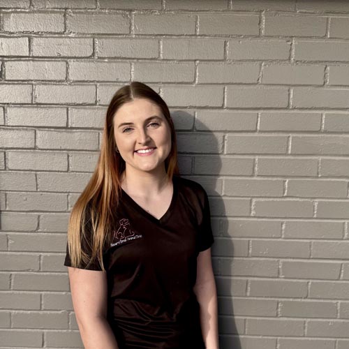 A woman with long hair stands against a gray brick wall, smiling and wearing a black shirt.