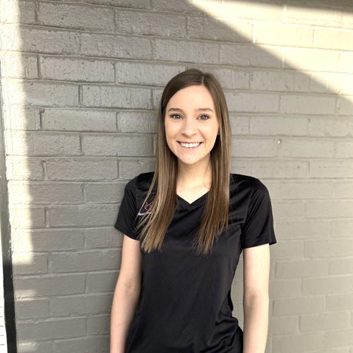 A woman with long, straight hair wearing a black shirt stands in front of a light gray brick wall, smiling at the camera.