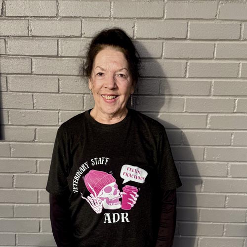 Woman standing in front of a gray brick wall wearing a black T-shirt with "Veterinary Staff ADR" and a skull design.