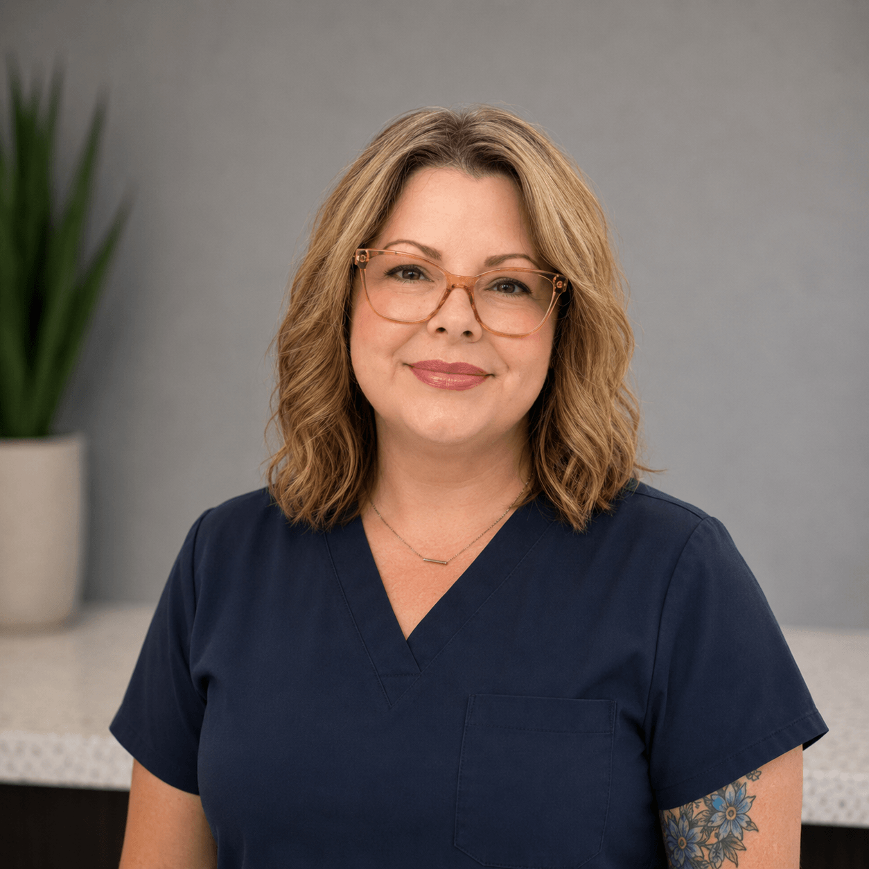 Woman with shoulder-length wavy hair and glasses wearing navy blue scrubs stands indoors, smiling. A plant in a pot is visible in the background.