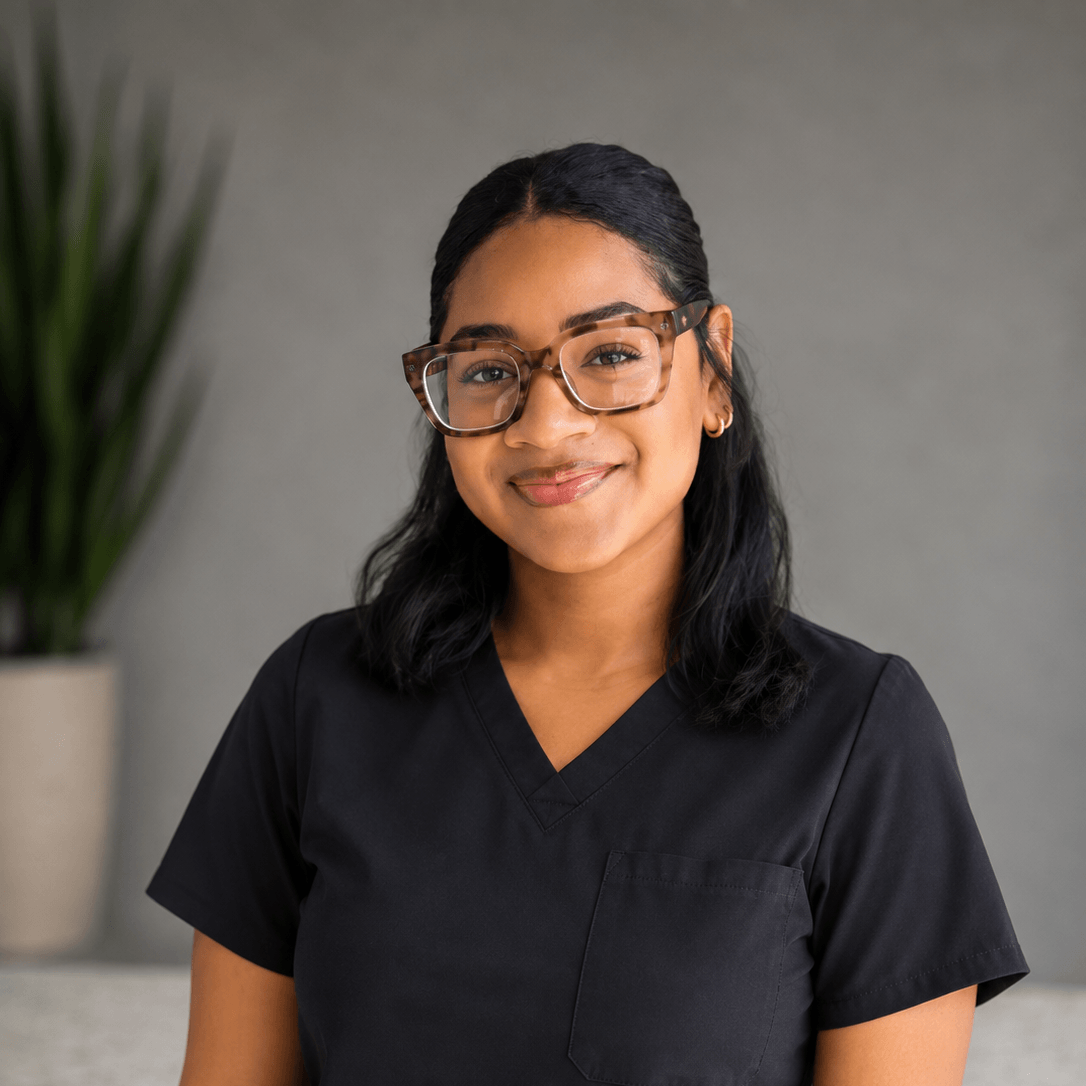 Woman wearing glasses and black scrubs smiles at the camera, standing indoors with a blurred plant in the background.