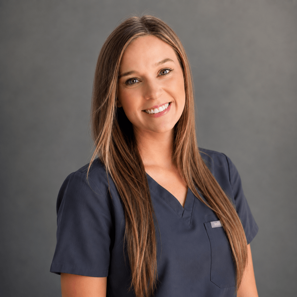 A woman with long straight hair, wearing navy blue medical scrubs, stands against a plain gray background and smiles at the camera.