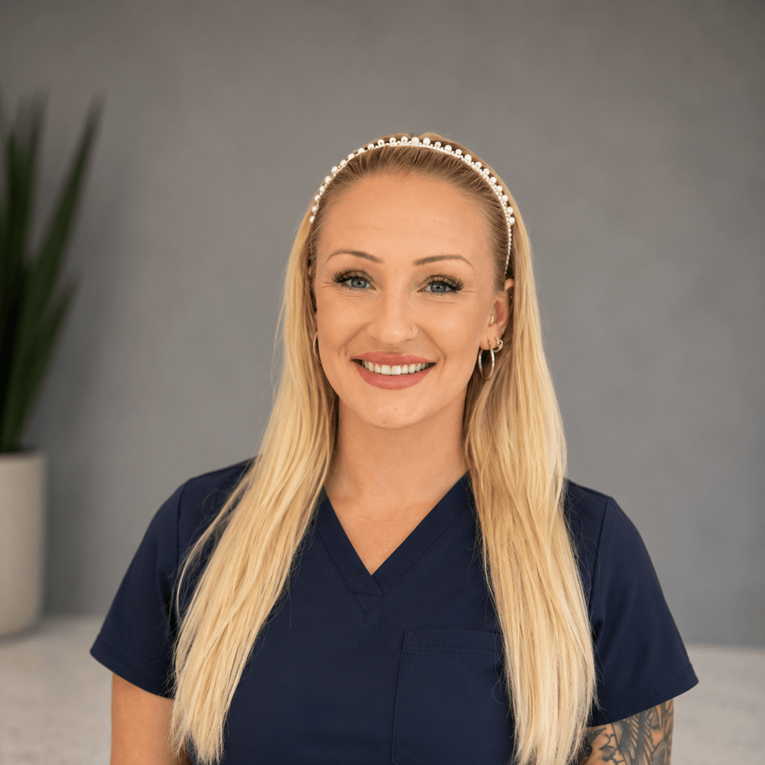 A woman with long blonde hair, wearing navy blue scrubs and a pearl headband, smiles at the camera. A potted plant is visible in the blurred background.